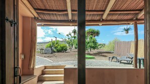 A view from a room with a stone floor and a wooden ceiling, looking out to a patio with a wooden fence, a tree, and a bench.
