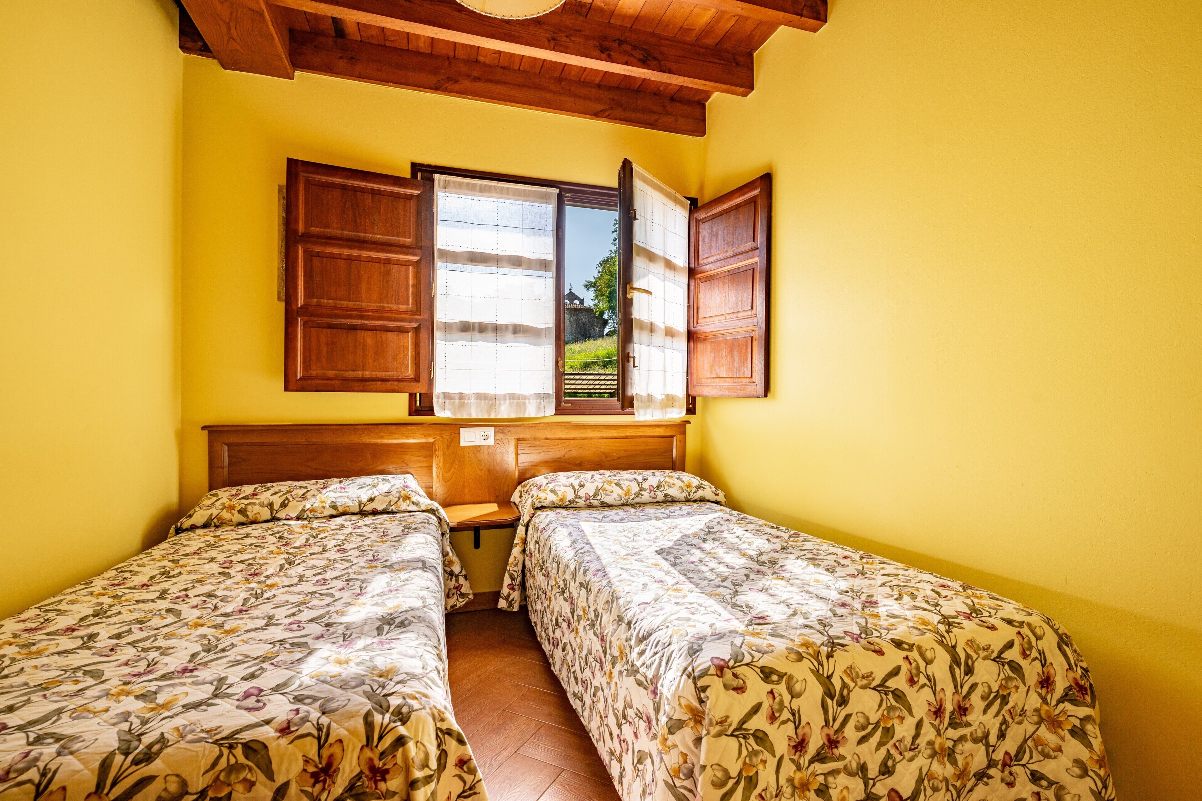 Habitación con dos camas, cabeceros de madera, una ventana con contraventanas abiertas y vistas al exterior con vegetación.