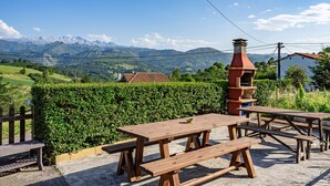 Ein Picknickbereich mit Holzbänken und -tischen, einem Backsteinofen und Blick auf Berge sowie ein Dorf.