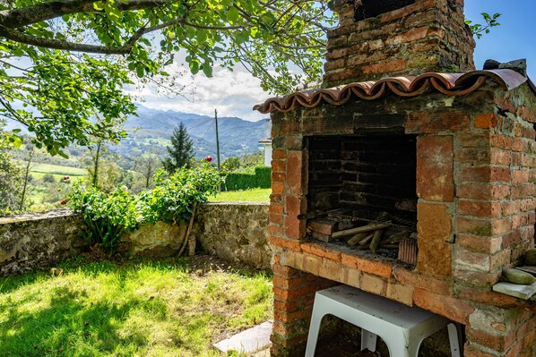 A brick-built outdoor oven with a white stool in front, surrounded by greenery and a mountainous backdrop.