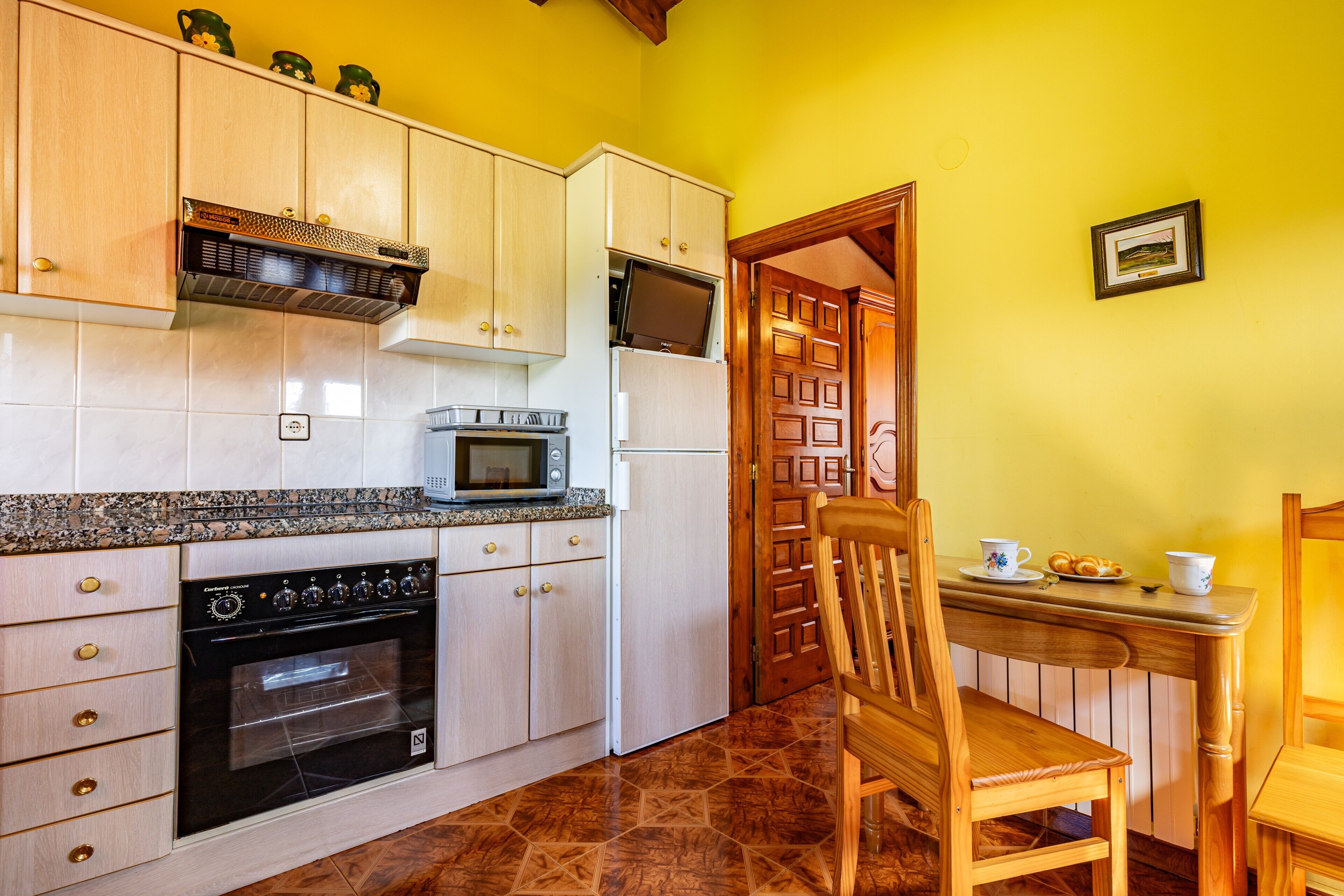 A kitchen with wooden cabinets, a microwave, a refrigerator, and a dining area with a chair and table.