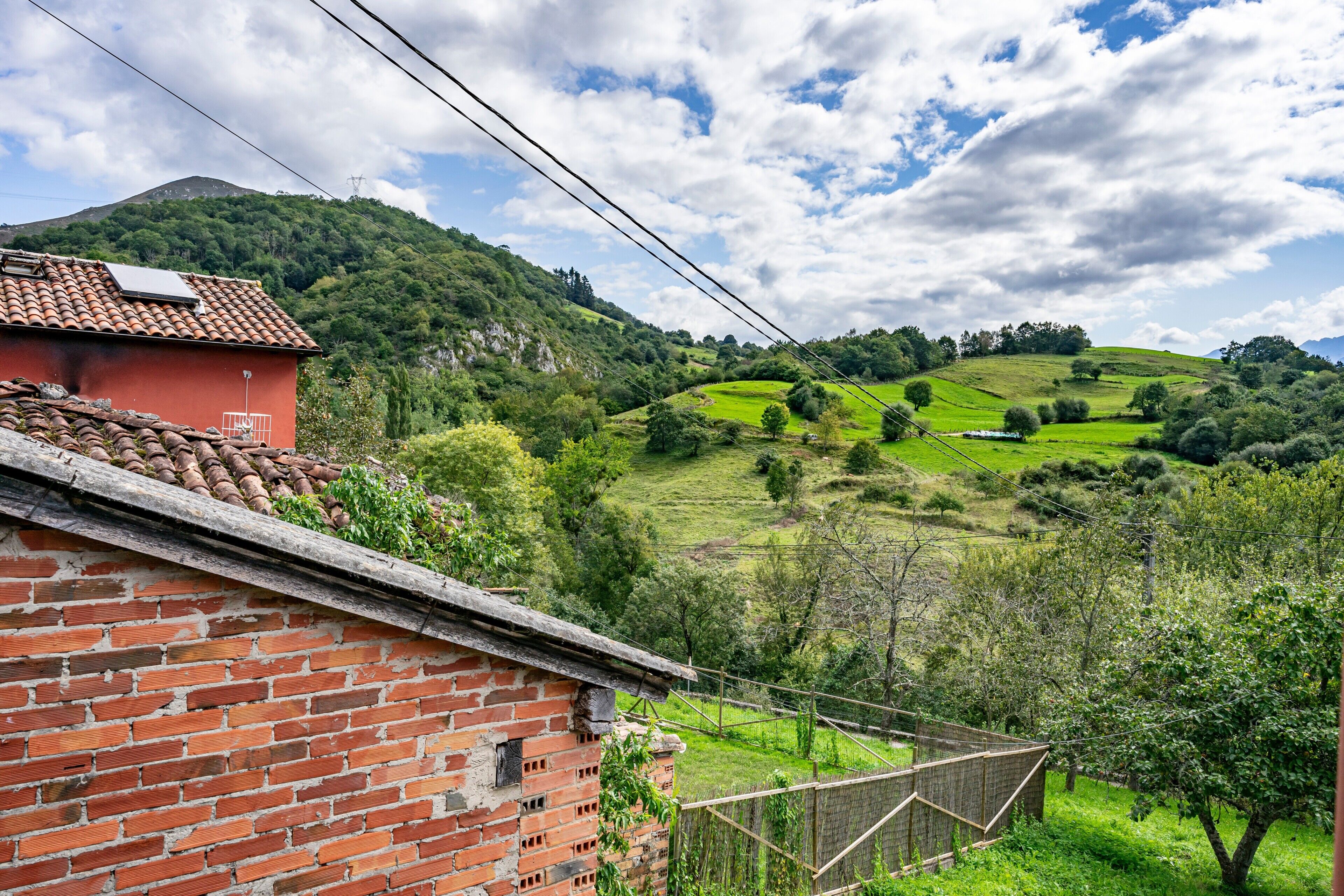 A rural landscape with a brick building, a fenced area, and rolling green hills under a partly cloudy sky.