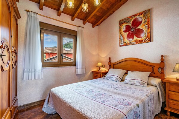 Un dormitorio con cama de madera, una ventana con cortinas, una mesita de noche con lámpara y un cuadro en la pared.