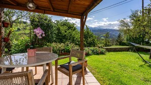 Eine Terrasse im Freien mit einem runden Glastisch, Korbstühlen und Blick auf einen üppigen Garten und Berge.