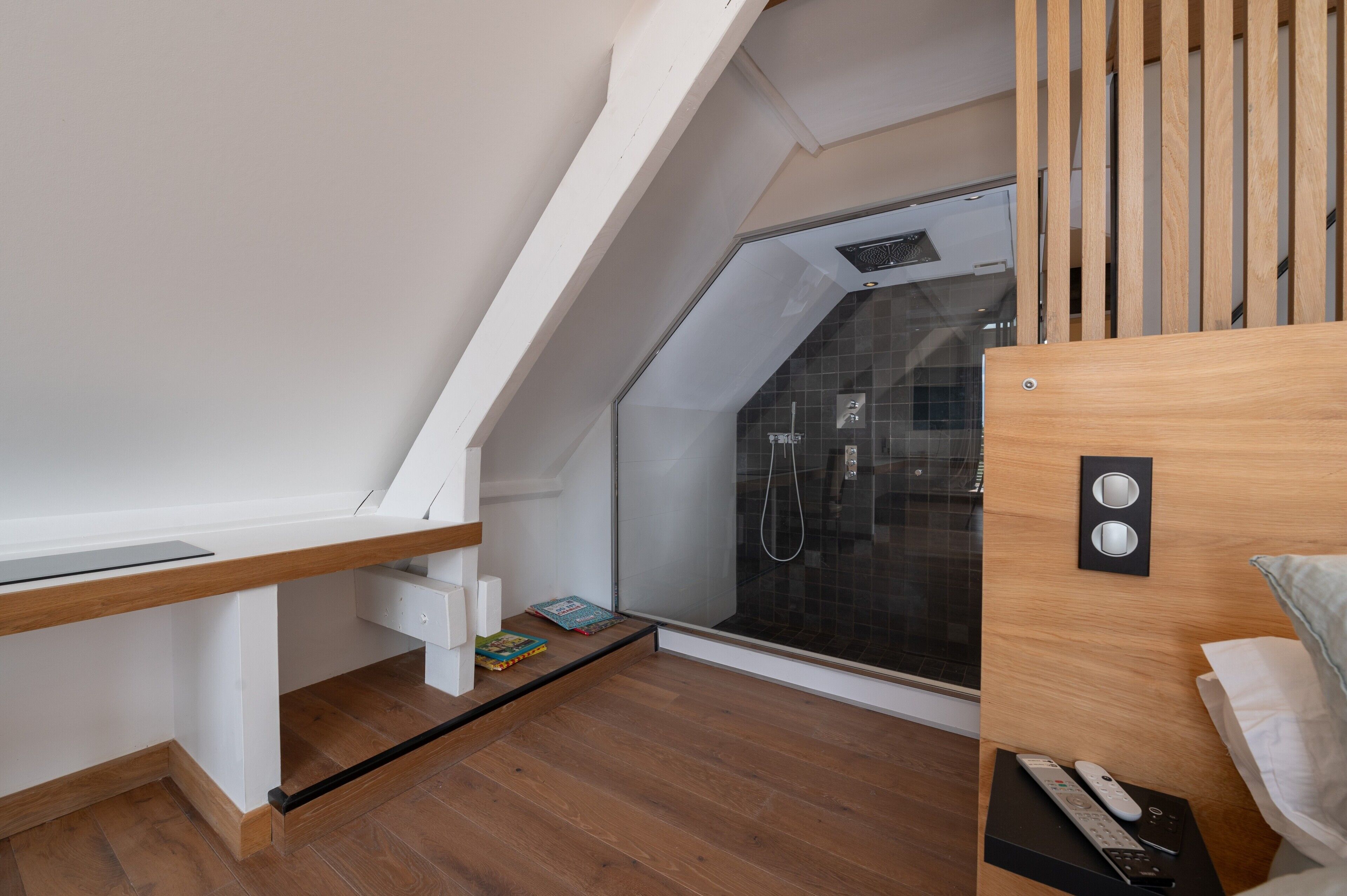 Une chambre moderne avec un plafond en pente, une cabine de douche en verre, un bureau en bois et un lit doté d’une tête de lit.