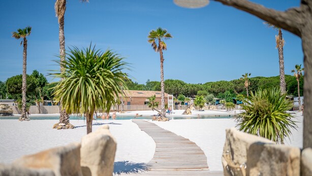 A wooden boardwalk leading to a beach area with palm trees and a building in the background. - Caravan 'Valras Plage 5 Étoiles' with Shared Pool, Wi-Fi and Air Conditioning (Vendres)