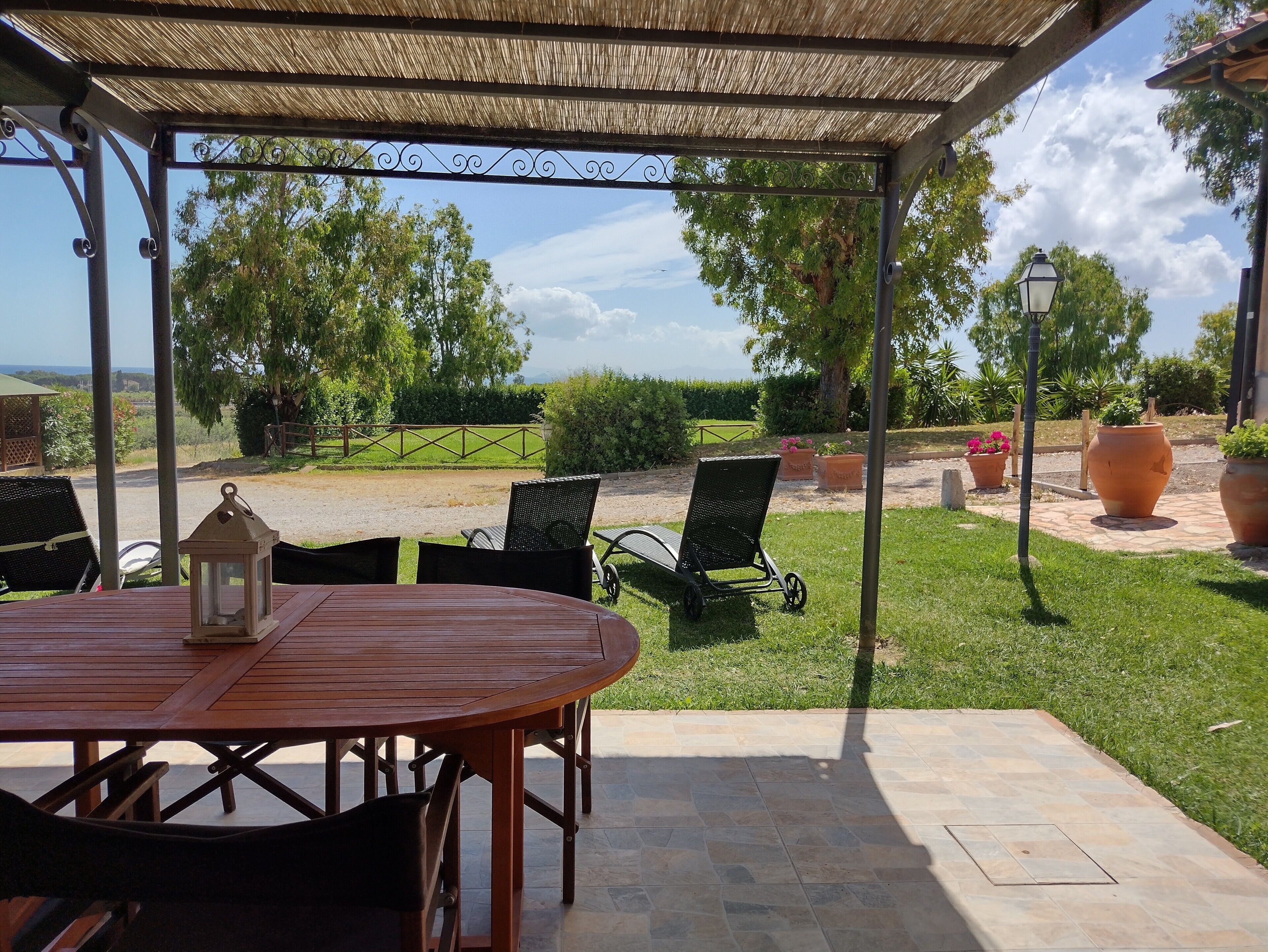 A covered patio with a table and chairs, a lantern, and a view of a landscaped garden with a fence and trees.