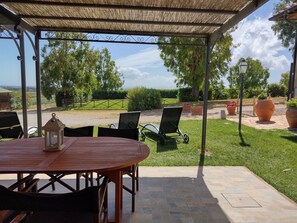 A covered patio with a table and chairs, a lantern, and a view of a landscaped garden with a fence and trees.