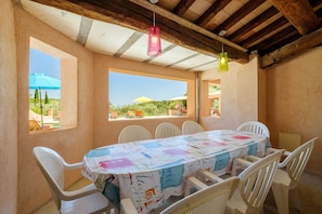Un coin repas avec une table recouverte d’une nappe colorée, des chaises blanches et une vue sur l’extérieur.