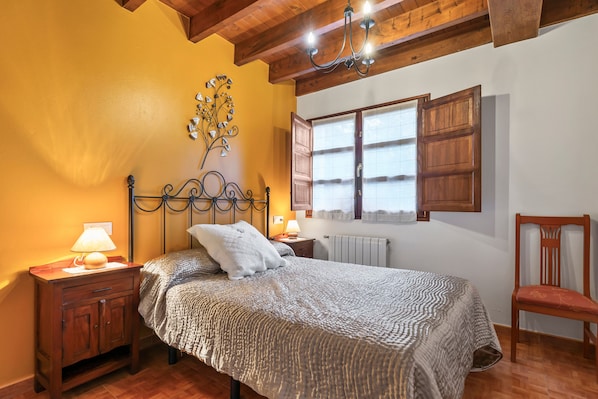 A bedroom with a bed, wooden nightstand, lamp, decorative wall piece, window with shutters, and a chair.