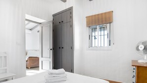 Une chambre à coucher comprenant un lit, une armoire, une fenêtre avec des stores, un ventilateur et donnant sur une autre pièce.