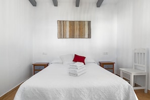 A neatly made bed with white linens, a red pillow, and a folded towel stack. There is a white chair and a small wooden nightstand with a drawer.