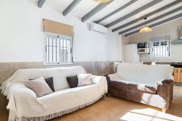 A living room with two sofas, a kitchen area, and a window with blinds.
