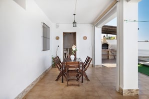 A dining area with a wooden table and chairs, a white vase with flowers, and a view of an outdoor patio with a barbecue.