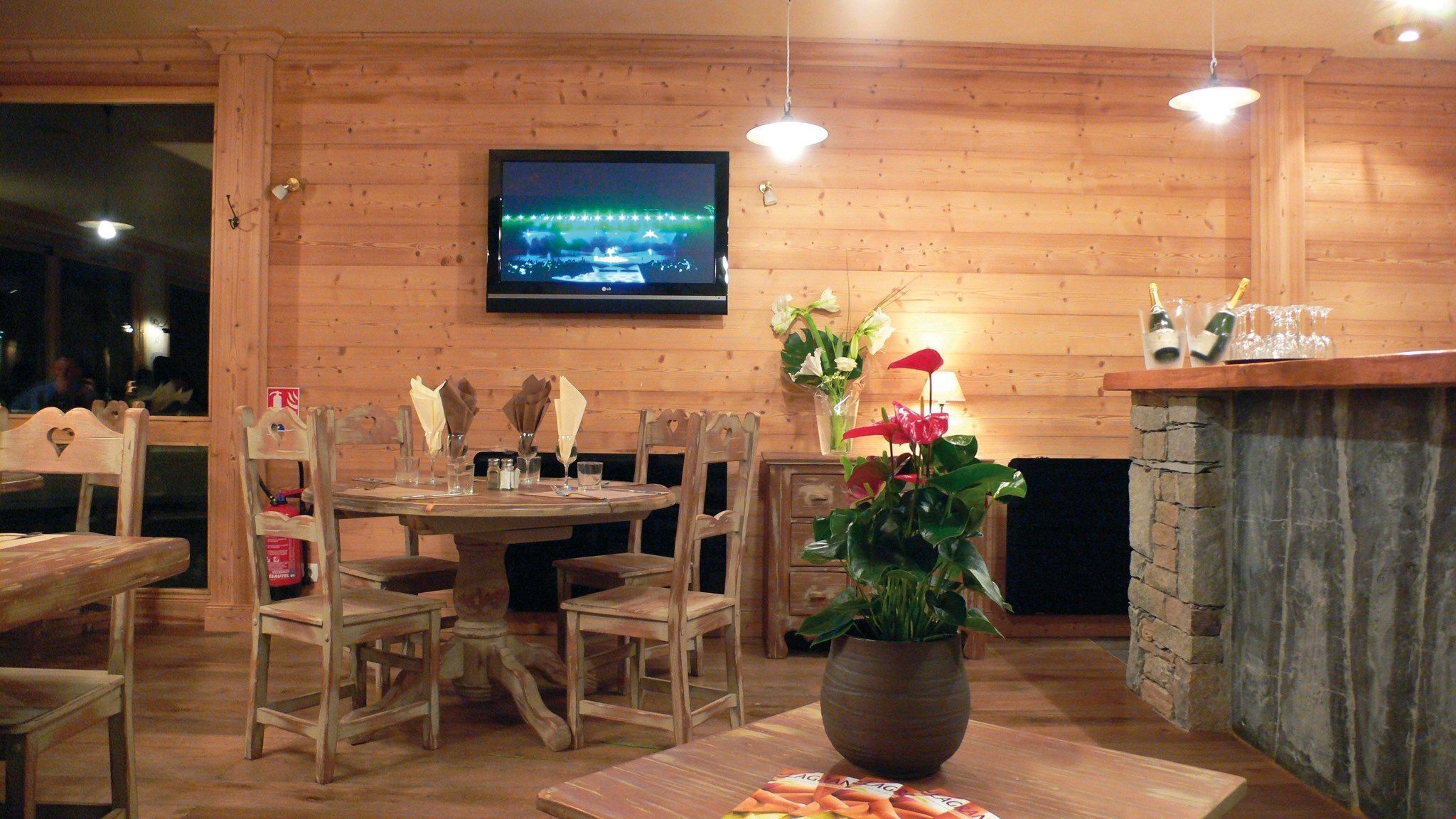 Un interior de madera con una mesa redonda para comer, sillas, un televisor montado en la pared y una chimenea de piedra.