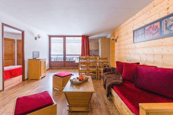 A cozy room with a wooden floor, a red sofa, a wooden table with fruit, and a view of a balcony.