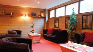 Ein gemütlicher Raum mit Holzwänden, einem roten Teppich und bequemen Sitzgelegenheiten, darunter eine Couch und Sessel. Es gibt Regale mit Büchern und Dekoration, sowie ein Fenster mit Tageslicht.