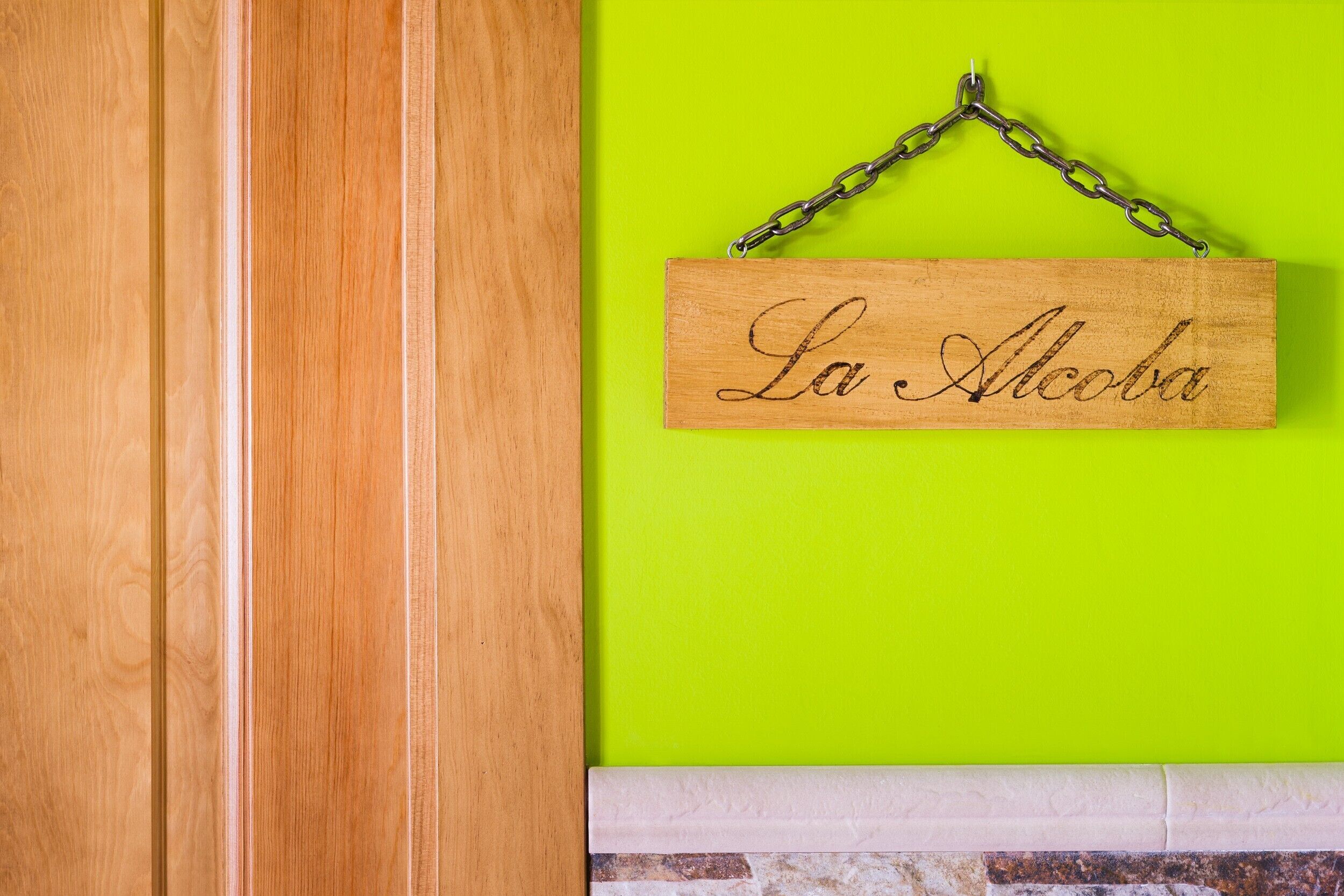 Un letrero de madera con 'La Alcoba' colgado en una pared verde brillante.