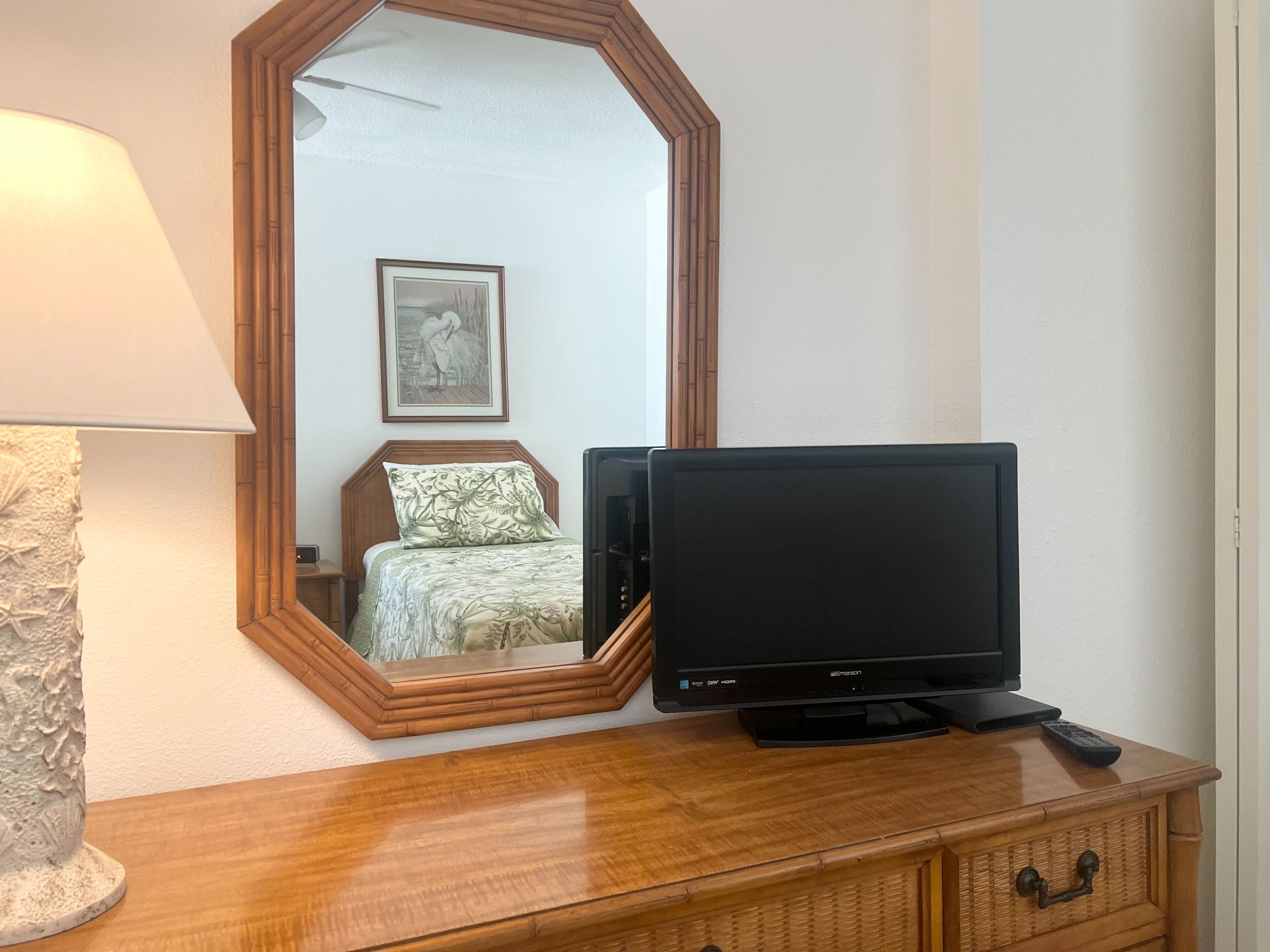 A wooden dresser with a flat-screen TV, a lamp, and a mirror reflecting a bed and a framed picture on the wall.