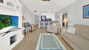 Una sala de estar moderna con una televisión de pantalla plana, un sofá beige, una mesa de centro y una cocina al fondo.