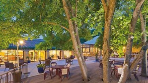 Een terras met houten tafels en stoelen, omringd door bomen en een gebouw op de achtergrond.