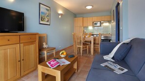 Una sala de estar con televisor, muebles de madera, una mesa de comedor con sillas y una cocina al fondo.