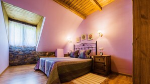 A bedroom with a stone wall, wooden ceiling beams, a bed with a patterned bedspread, a nightstand with a lamp, and a wooden wardrobe. - Casa Rural Devesa in Villablino, Laciana Valley with Pool (Caboalles De Abajo)