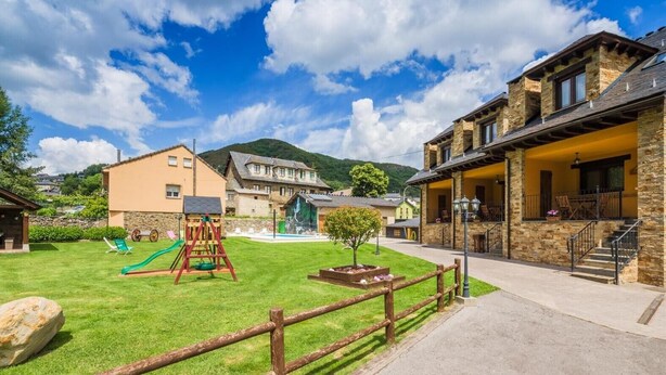 A courtyard with a swing set, a small tree, and a stone building with a balcony. - Casa Rural Devesa in Villablino, Laciana Valley with Pool (Caboalles De Abajo)