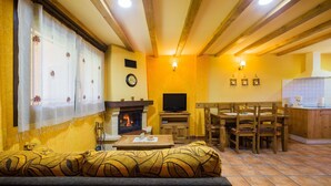 A cozy living room with a fireplace, wooden furniture, and a dining area with a kitchen in the background. - Casa Rural Devesa in Villablino, Laciana Valley with Pool (Caboalles De Abajo)