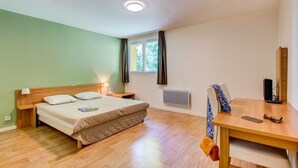 Un dormitorio con cama, mesita de noche, lámpara, ventana, radiador y un escritorio con silla.