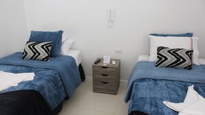 Une chambre d’hôtel avec deux lits, chacun recouvert d’une couverture bleue et orné d’un oreiller à motifs noir et blanc. Une table de chevet en bois, sur laquelle se trouvent un téléphone et une feuille de papier, est placée entre les deux lits.