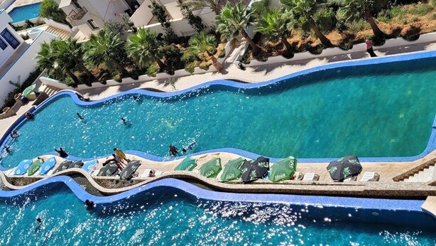 A large, winding pool with clear blue water, surrounded by palm trees and a white building. - CTM - Le Bateau (Terga)