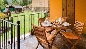 Un balcón con una mesa lista para el desayuno, una valla de madera y vistas a una casa y montañas.