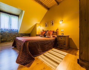 A bedroom with a sloped ceiling, a bed with a quilt, a nightstand, a wall-mounted lamp, and a stone wall. - Casa Rural Balagar in Villablino, Laciana Valley with Pool (Caboalles De Abajo)
