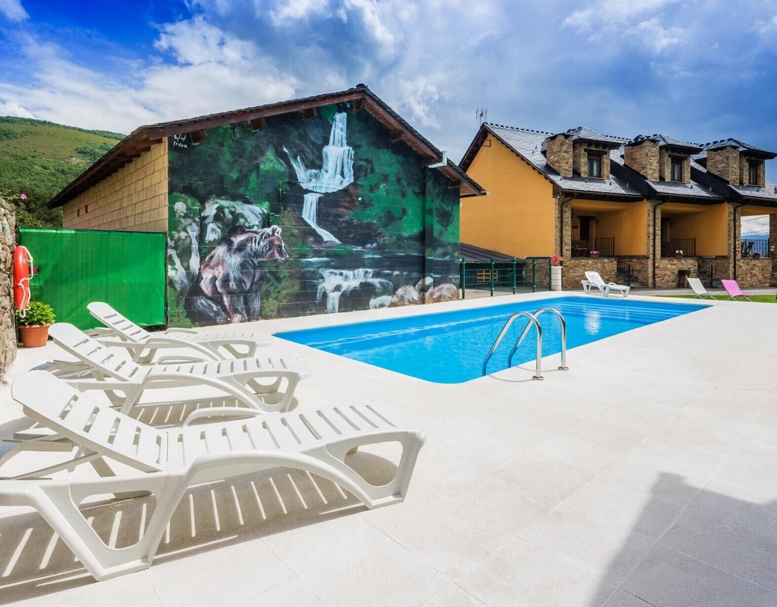 Una piscina con sillas de descanso, un mural con una cascada y un oso, y un edificio de piedra con un porche cubierto.