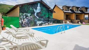 Una piscina con sillas de descanso, un mural con una cascada y un oso, y un edificio de piedra con un porche cubierto.