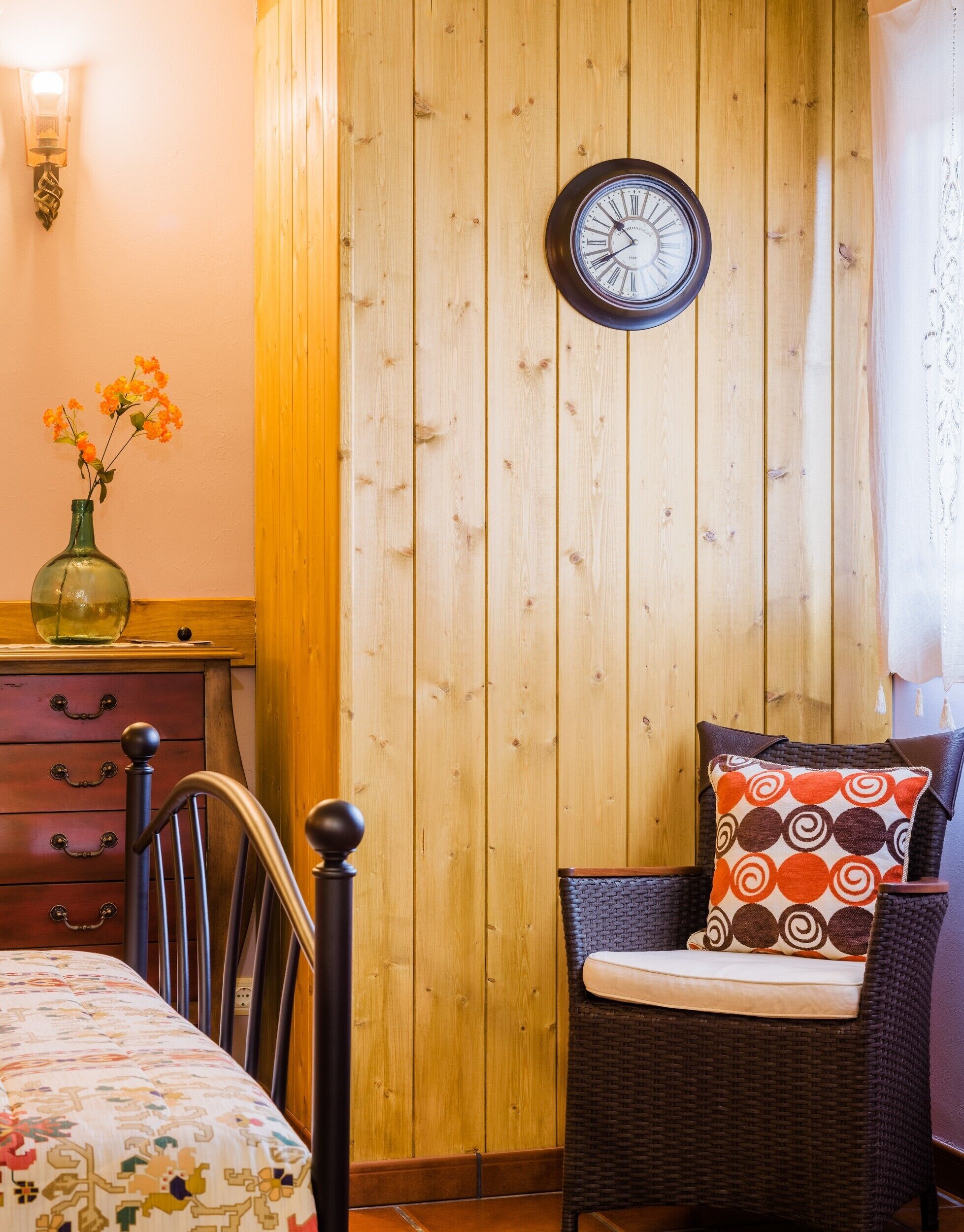 Una habitación con paneles de madera, una cama, una silla con cojín estampado, un reloj de pared y un florero con flores.