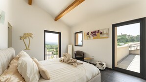 A bedroom with a large bed, a chair, a small table, and a balcony with a view.