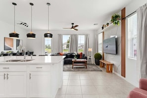 A modern kitchen with a central island, a dining area with a sofa and coffee table, a living room with a TV, and a ceiling fan.