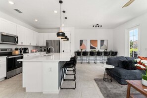 Une cuisine moderne avec des meubles blancs, un îlot central, des appareils électroménagers en acier inoxydable et un coin repas avec des chaises noires et un canapé gris.