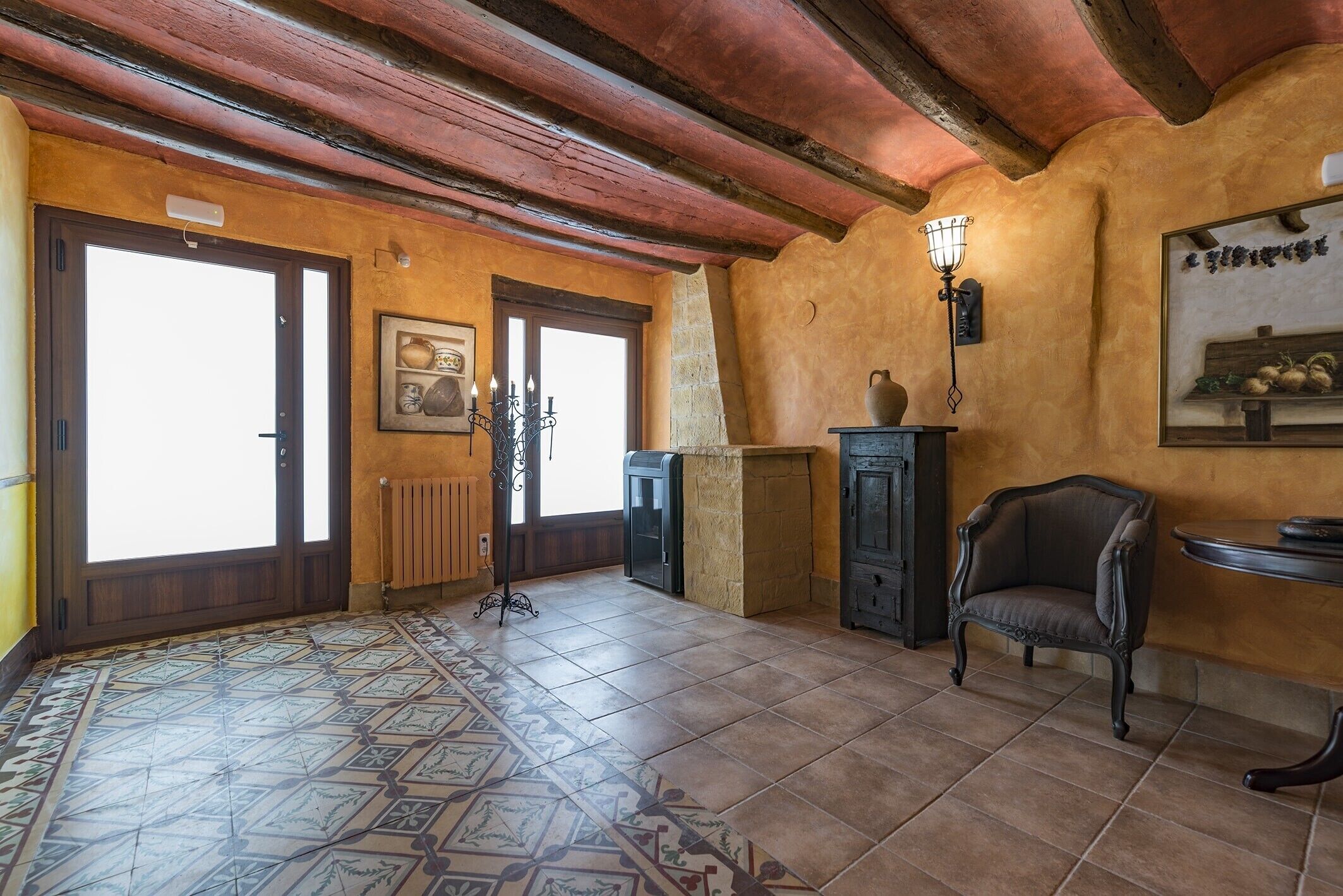 Una habitación con piso de baldosas, vigas de madera en el techo, una silla, una mesa y un aplique de pared.
