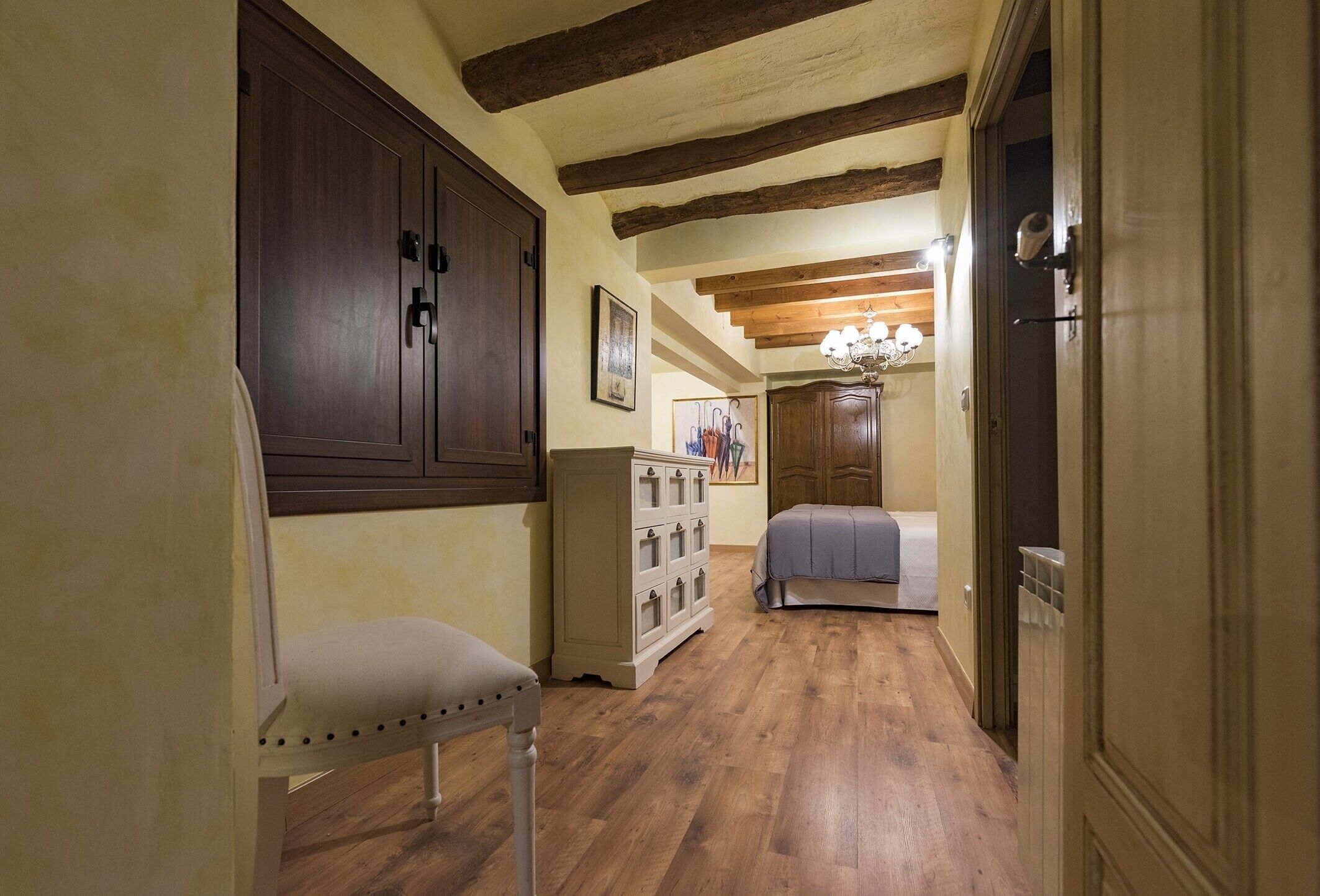 A hallway with wooden flooring, a large dark wooden wardrobe, a white upholstered chair, a white dresser, and a bed with a gray bedspread.