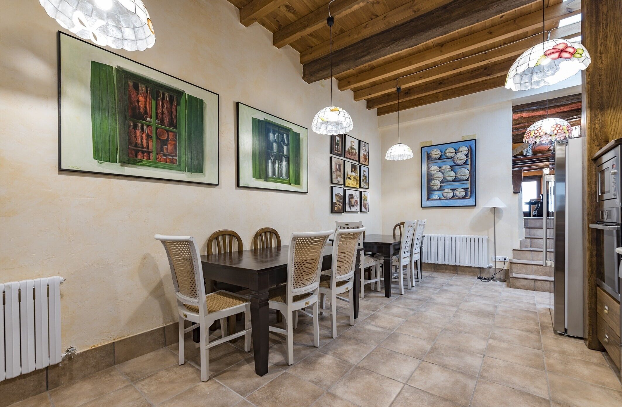A dining area with a wooden ceiling, tiled floor, and a white radiator. There are multiple dining tables and chairs, and wall-mounted light fixtures.