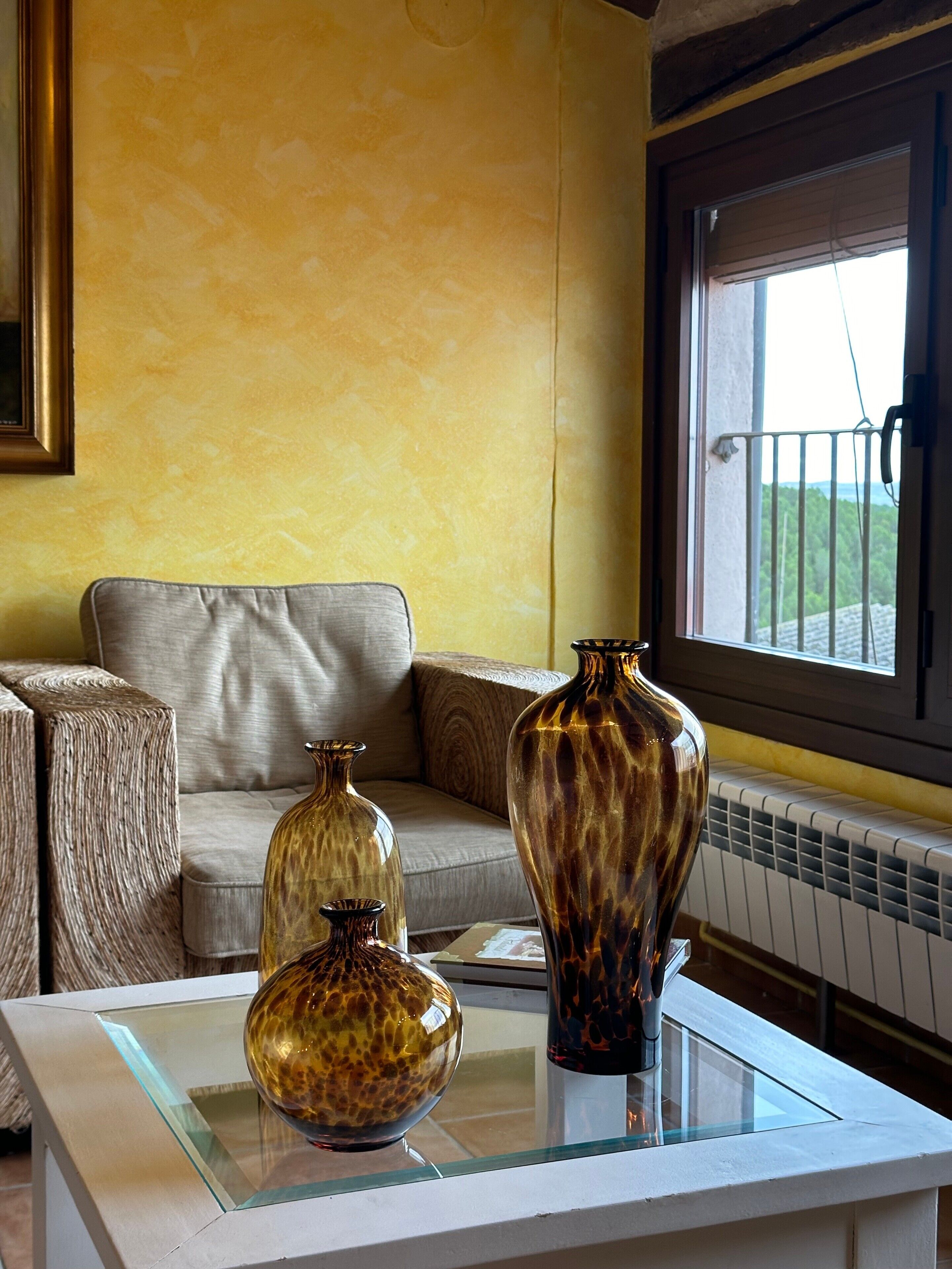 A room with a sofa, two glass-top tables with decorative vases, a window with a view, and a radiator.
