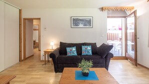 Een woonkamer met een bank, een bijzettafeltje met een plant, een ingelijst schilderij aan de muur en een deur naar een buitenruimte.