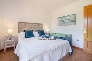 A neatly made bed with a white comforter, blue pillows, and a green blanket. There is a painting of a beach scene on the wall, a small bedside table with a lamp, and a wooden door.