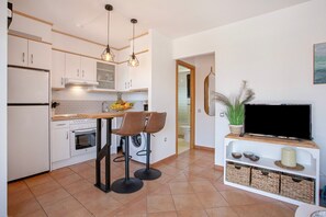 A modern kitchen with white cabinets, a built-in oven, a dining area with two stools, and a TV on a wooden stand.