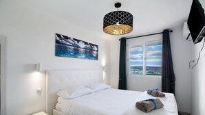 A hotel room with a bed, a nightstand, a wall-mounted TV, a window with curtains, and a painting of a seascape. - Vacation Apartment 'Francesco House 1509 Monte Paraiso' with Sea View, Wi-Fi and Air Conditioning (Puerto Rico de Gran Canaria)