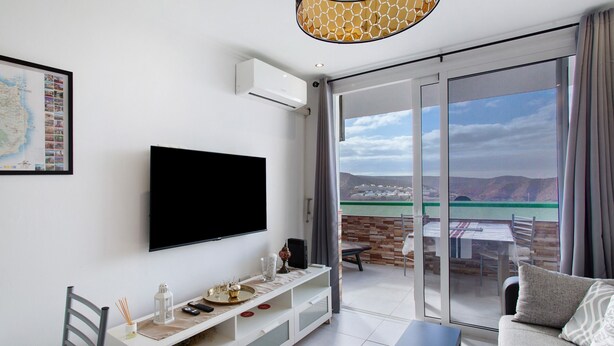 A modern living room with a flat-screen TV, a white entertainment unit, a sofa, and a sliding glass door leading to an outdoor patio with a table and chairs. - Vacation Apartment 'Francesco House 1509 Monte Paraiso' with Sea View, Wi-Fi and Air Conditioning (Puerto Rico de Gran Canaria)