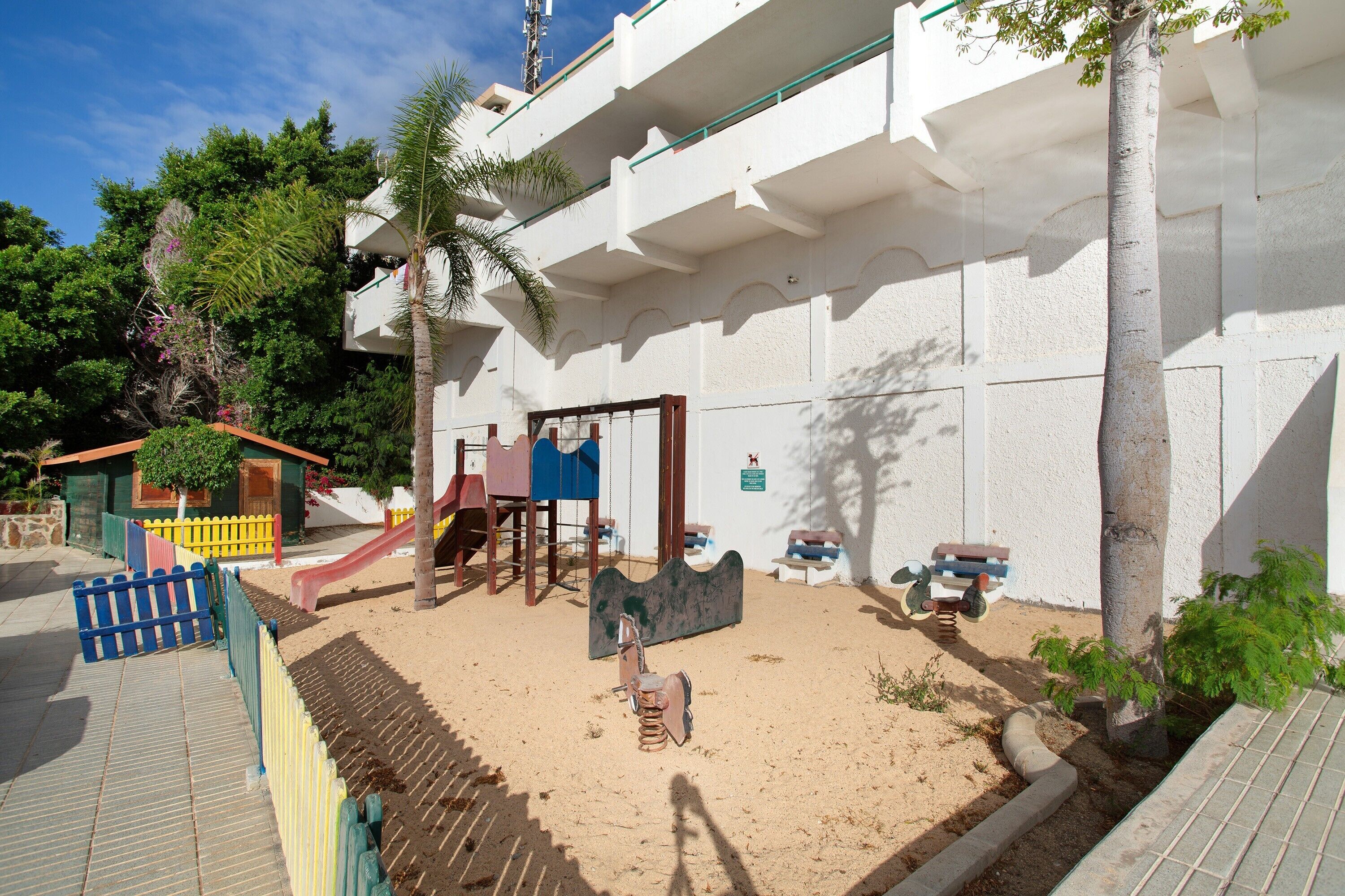 Un terrain de jeux avec un toboggan et des balançoires, un petit bâtiment et un palmier.